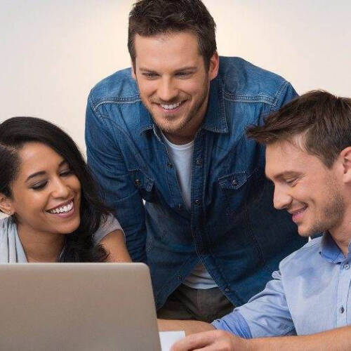 Three people, two men and a woman look at a laptop smiling in what seems like an information sharing moment. On of the men in denim jacket is standing.